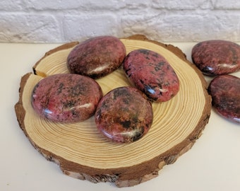May include: Five smooth, polished pink and black stones with a marbled pattern. The stones are arranged on a wooden slice.