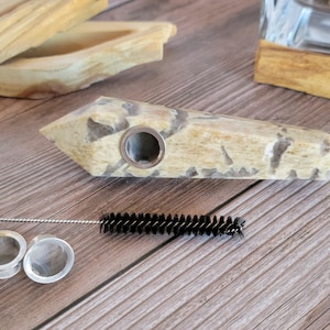 May include: A light brown and white crystal pipe with a round metal bowl. The pipe is on a wooden surface with a small brush and two metal screens.
