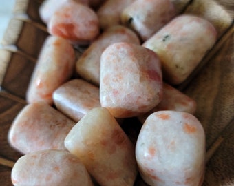 Puede incluir: Una colección de cristales de piedra solar pulidos en un cuenco de madera. Los cristales son una mezcla de naranja claro y blanco con una calidad brillante e iridiscente.