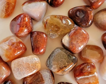 May include: A collection of carnelian tumbled stones in various shades of orange, brown, and white. The stones are smooth and polished, with a variety of patterns and textures.
