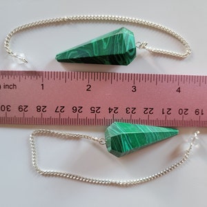 May include: Two green malachite crystal pendulums with silver chains, measured against a ruler. The pendulums are approximately 2 inches long.