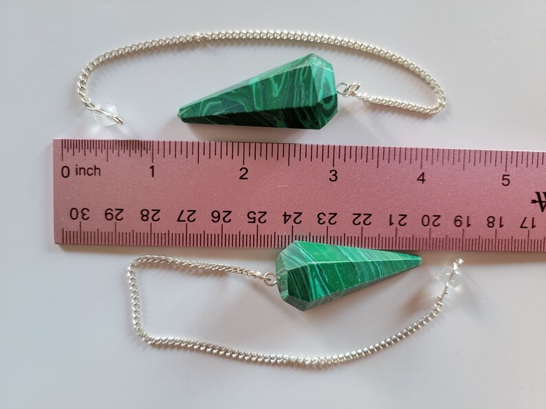 May include: Two green malachite crystal pendulums with silver chains, measured against a ruler. The pendulums are approximately 2 inches long.