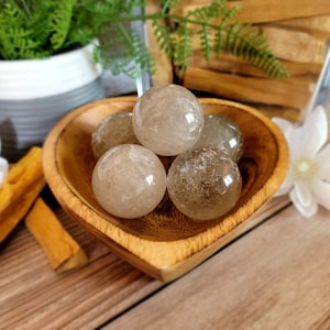 May include: Four clear quartz crystal spheres in a wooden bowl. The spheres are translucent and have a cloudy appearance. The bowl is made of light brown wood and has a heart shape.