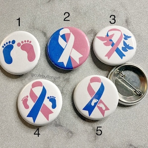Puede incluir: Conjunto de seis botones de 2,5 cm de diámetro con varios diseños. Los diseños incluyen una cinta azul y rosa, un par de huellas de pies de bebé y un par de palomas. Los botones son blancos con un esquema de color azul y rosa.