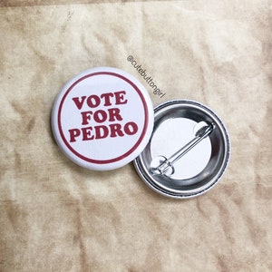 Puede incluir: Un botón blanco con un borde rojo y el texto "VOTE FOR PEDRO" en letras rojas. La parte posterior del botón es plateada con un alfiler.