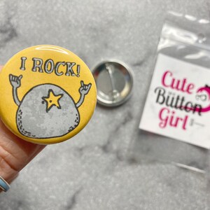 Peut inclure: Un bouton jaune avec un rocher de dessin animé avec une étoile dessus et le texte "I ROCK!"