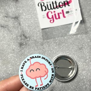Peut inclure: Un badge rond avec un fond bleu clair représentant un cerveau rose de dessin animé avec un visage souriant. Le texte sur le bord indique I HAVE A BRAIN INJURY PLEASE BE PATIENT WITH ME. Un deuxième badge est visible à proximité.