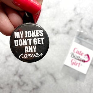 May include: A black button with white text that reads "My jokes don't get any cornea". The button is held by a hand with red nail polish. The button is packaged in a clear plastic bag with a white label that reads "Cute Button Girl".