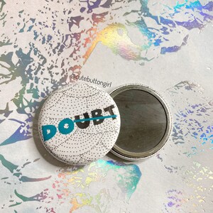 May include: A white button with a black and blue design that says "DOUBT" with a line through it. The button is attached to a silver mirror.