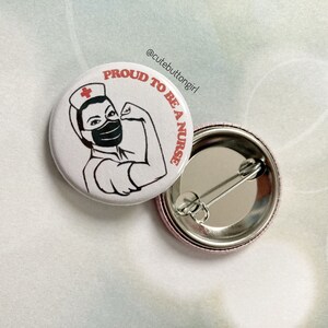 Könnte beinhalten: Weißer Knopf mit einer Schwarzweißillustration einer Krankenschwester, die eine Gesichtsmaske und eine rote Krankenschwestermütze trägt. Der Text "PROUD TO BE A NURSE" ist in Rot um die Illustration herum geschrieben. Die Rückseite des Knopfes ist silberfarben mit einer Sicherheitsnadel.