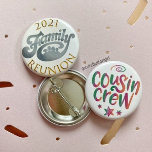 Peut inclure: Deux épingles à bouton blanches avec du texte coloré. La première épingle dit "2021 Family Reunion" en lettres dorées. La deuxième épingle dit "Cousin Crew" en lettres arc-en-ciel avec des étoiles.