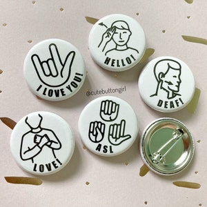 Può includere: Sei bottoni rotondi bianchi con contorni neri e testo. I bottoni presentano diversi simboli della lingua dei segni e testo, tra cui "I Love You!" "Hello!" "Deaf!" "Love!" "ASL" e un bottone vuoto con uno spillo.