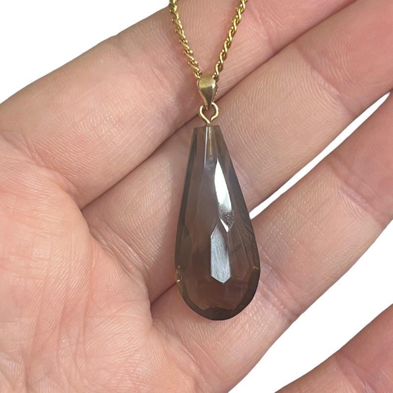 May include: A gold-colored chain necklace with a teardrop-shaped pendant. The pendant is a faceted, smoky-brown gemstone. The necklace is held in a person's hand against a white background. Jewelry.