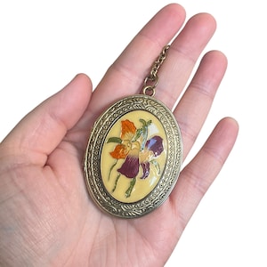 May include: Oval-shaped pendant with a floral design. The pendant features a cream-colored background with orange, purple, and white flowers. The pendant has a gold-colored metal frame and a small chain.