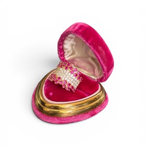 May include: A wide gold band ring featuring multiple rows of round, vibrant pink stones and a central row of clear, sparkling white stones, presented inside a heart-shaped, fuchsia-colored velvet jewelry box with a gold-toned metal trim.