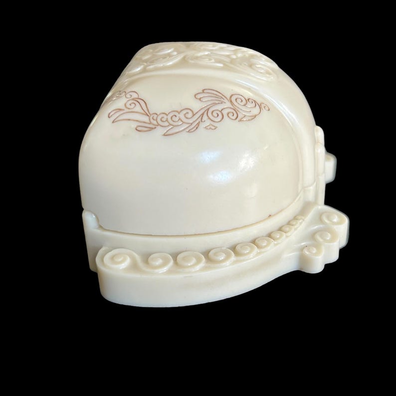 K&ouml;nnte beinhalten: Eine kleine, cremefarbene Ringbox mit einem dekorativen, verzierten Design. Die Box hat einen Klappdeckel und ein Blumenmuster.