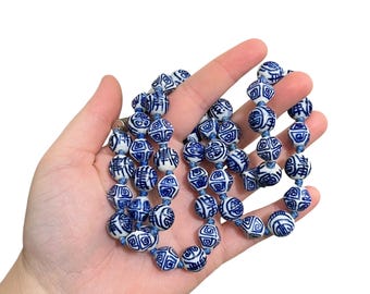 May include: A necklace of white and blue ceramic beads with intricate patterns. The beads are strung on a blue cord and held in a hand against a white background. The beads are round and have a glossy finish.