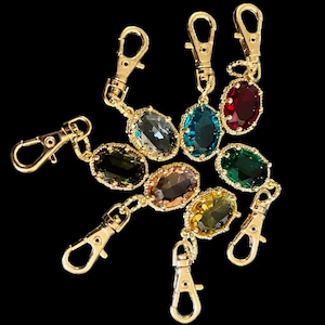 Puede incluir: Llaveros con cierre de mosquetón de metal dorado con colgantes de piedras preciosas ovaladas de colores. Las piedras preciosas son en tonos verdes, azules, rojos y amarillos. Los llaveros se muestran sobre un fondo negro.