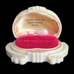 K&ouml;nnte beinhalten: Eine wei&szlig;e Plastik-Ringbox mit rotem Samt-Innenfutter. Die Box ist mit "MITCHELL-COPP, LTD. Diamond Merchants Winnipeg." beschriftet.