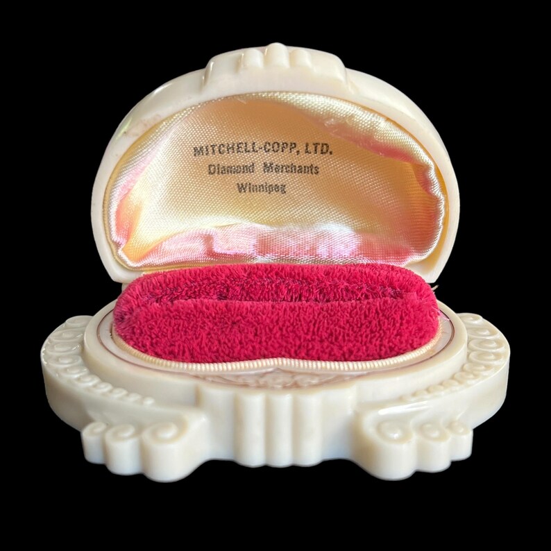 K&ouml;nnte beinhalten: Eine wei&szlig;e Plastik-Ringbox mit rotem Samt-Innenfutter. Die Box ist mit "MITCHELL-COPP, LTD. Diamond Merchants Winnipeg." beschriftet.