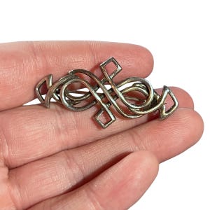 May include: Silver-toned brooch with an intricate, flowing design. The brooch features interwoven lines and geometric shapes, creating a complex and elegant pattern. The metal has a slightly aged appearance, adding to its vintage charm.