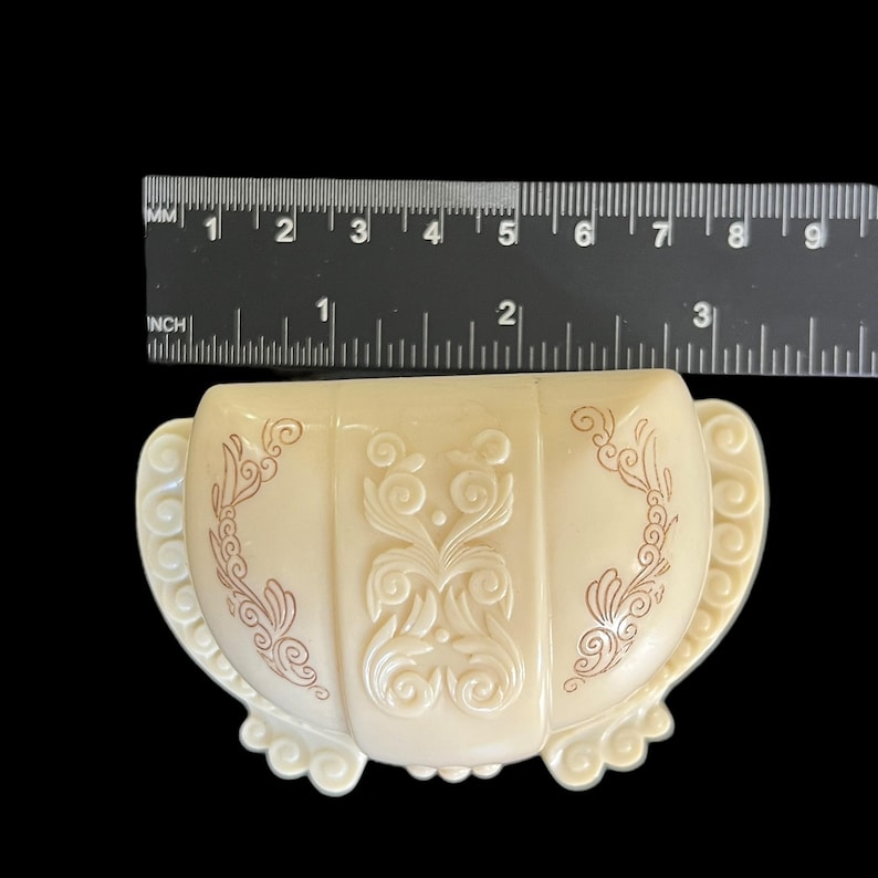 K&ouml;nnte beinhalten: Eine kleine, cremefarbene Schmuckschatulle mit einem dekorativen, erhabenen Design in rotbrauner Farbe. Die Schachtel ist muschelf&ouml;rmig und hat einen gewellten Rand.