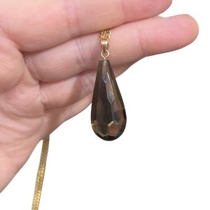 May include: A gold-colored chain necklace with a faceted, teardrop-shaped pendant. The pendant is a smoky, translucent brown color. The necklace is held in a person's hand against a white background.