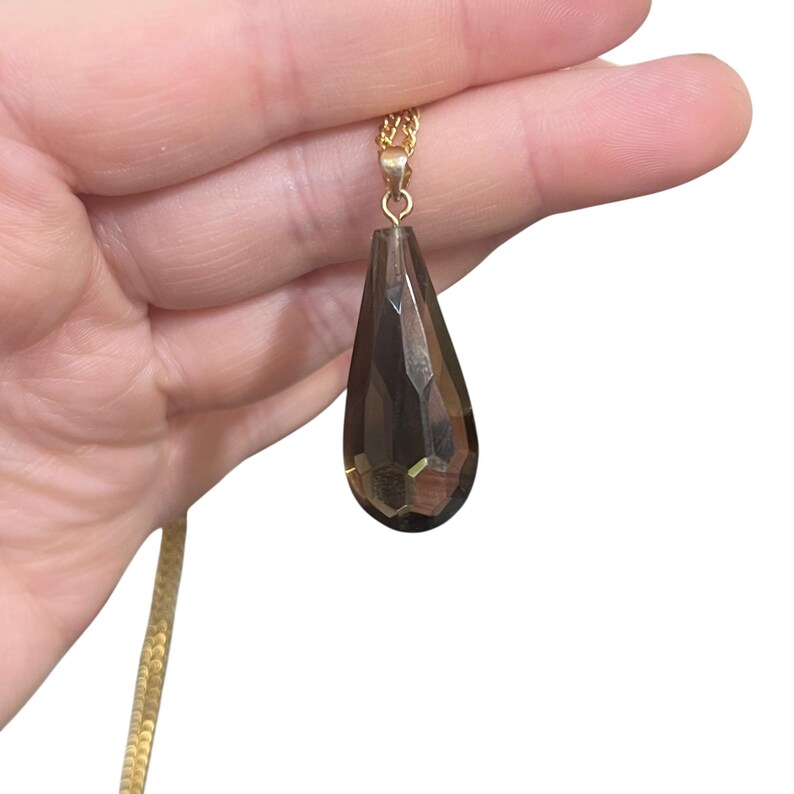 May include: A gold-colored chain necklace with a faceted, teardrop-shaped pendant. The pendant is a smoky, translucent brown color. The necklace is held in a person's hand against a white background.