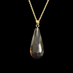 May include: A gold-colored chain necklace with a faceted, teardrop-shaped pendant. The pendant is a smoky, translucent color and hangs from a small gold bail. The background is solid black.