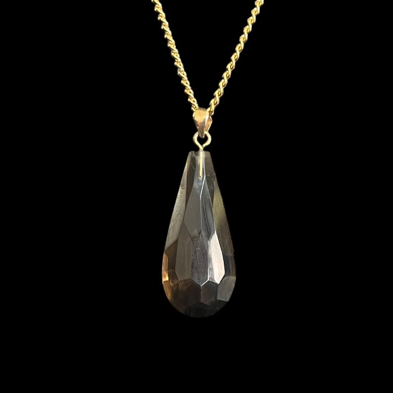 May include: A gold-colored chain necklace with a faceted, teardrop-shaped pendant. The pendant is a smoky, translucent color and hangs from a small gold bail. The background is solid black.