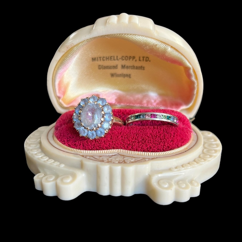 K&ouml;nnte beinhalten: Eine Vintage-Ringbox mit rotem Samt-Innenfutter. Die Box ist aus wei&szlig;em Kunststoff und hat ein dekoratives Design. Sie enth&auml;lt zwei Ringe, einen mit einem gro&szlig;en blauen Stein, der von kleineren blauen Steinen umgeben ist, und einen weiteren mit einer Reihe von bunten Steinen.