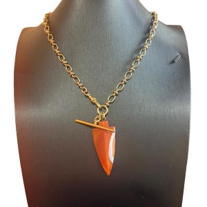 Op de afbeelding: Een goudkleurige ketting met een pijl-vormige hanger. De hanger is een mix van oranje en wit. De ketting wordt gepresenteerd op een zwarte mannequin. De ketting is gemaakt van kleine, verbonden schakels.