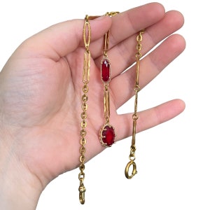 Könnte beinhalten: Eine goldene Kette mit zwei roten Edelstein-Akzenten. Die Kette hat einen Verschluss und ist zum Tragen als Uhrenkette gedacht.