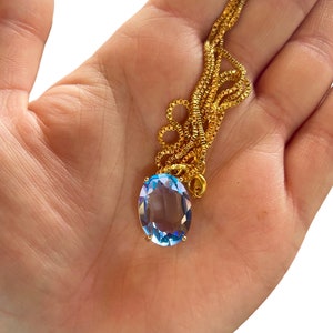 Op de afbeelding: Een gouden ketting met een lichtblauwe ovale edelsteen hanger.