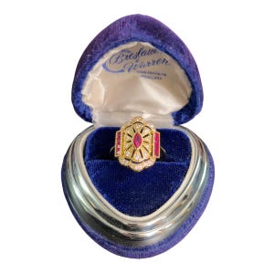 May include: A gold ring featuring a central oval ruby and rectangular rubies, encircled by small diamonds. The ring is presented in a blue velvet heart-shaped box, with the text "Breslau Warren" visible inside.