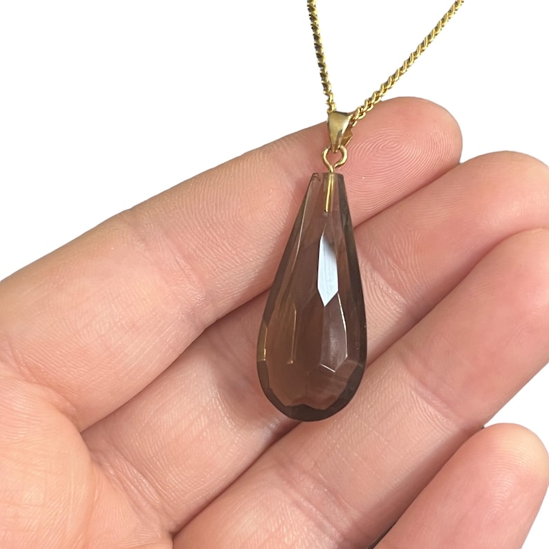 May include: A faceted, teardrop-shaped pendant necklace. The pendant is a translucent, smoky brown color and is suspended from a delicate gold chain. The necklace is held in a hand.
