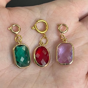 Puede incluir: Tres colgantes de amuleto de oro con piedras de vidrio facetadas de diferentes colores. Los amuletos son de forma rectangular con esquinas redondeadas. Las piedras son verdes, rojas y moradas.