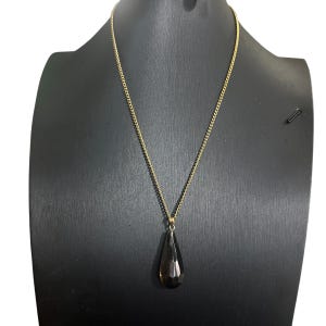 May include: A gold-colored chain necklace with a teardrop-shaped, dark brown pendant. The pendant is faceted, catching the light. The necklace is displayed on a black mannequin.