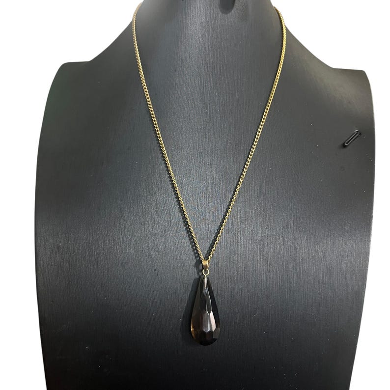 May include: A gold-colored chain necklace with a teardrop-shaped, dark brown pendant. The pendant is faceted, catching the light. The necklace is displayed on a black mannequin.