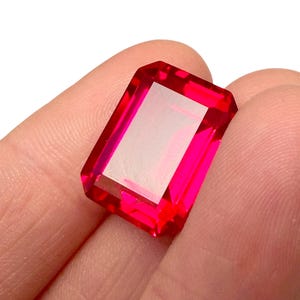 May include: A faceted, rectangular-cut, vibrant red gemstone. The gem is held between fingers, showcasing its bright color and sharp edges. The gem's cut allows light to reflect, creating a sparkling effect. The gemstone is likely for jewelry making.