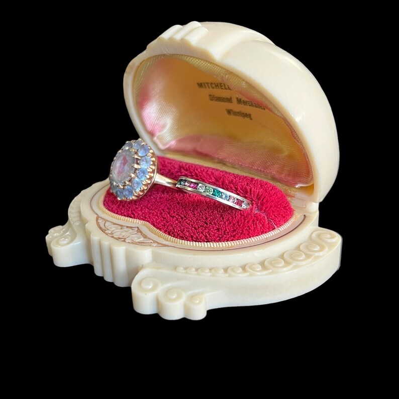 K&ouml;nnte beinhalten: Ein Ring mit einem gro&szlig;en blauen Edelstein, umgeben von kleineren blauen Edelsteinen, gefasst in einer goldenen Fassung. Der Ring befindet sich in einer wei&szlig;en Ringbox mit rotem Samt-Innenfutter. Die Box tr&auml;gt innen den Schriftzug "MITCHELL Diamond Merchants Winnipeg".
