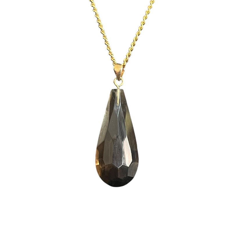 May include: A gold-colored chain necklace with a faceted, teardrop-shaped pendant. The pendant is a dark, smoky gray color and has a geometric design. The necklace is displayed against a white background.