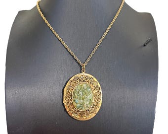 Puede incluir: Un collar con colgante dorado con un centro ovalado texturizado lleno de piedras verdes y azules. El colgante está rodeado por un diseño de filigrana ornamentado y cuelga de una delicada cadena.