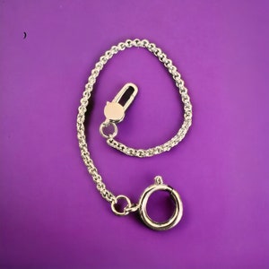 May include: A silver-toned chain with a lobster clasp and a large circular ring. The chain is made of small, interconnected links. The clasp and ring are shiny and reflective, set against a purple background. The chain is likely for a bag or accessory.