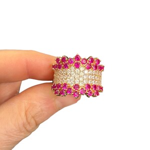 May include: A gold-toned ring featuring a wide band design. The centre section is densely set with multiple rows of small, clear, round-cut stones, bordered on the top and bottom by a single row of vibrant, round-cut pink stones in a scalloped edge.