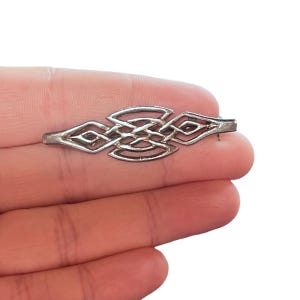 May include: A silver-toned brooch with an intricate Celtic knot design. The brooch features a symmetrical pattern of interwoven lines and diamond shapes. The pin is held in place by a clasp on the back.