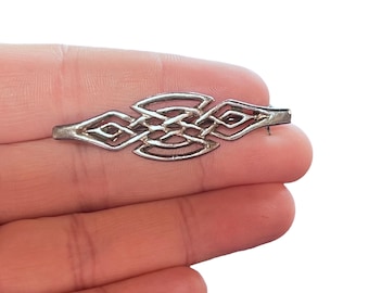 Pode incluir: Um broche prateado com um intrincado design de nó celta. O broche apresenta um padrão simétrico de linhas entrelaçadas e formas de diamante. O alfinete é preso por um fecho na parte de trás.