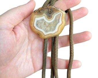 Può includere: Un bolo tie in agata marrone e bianca con un cordoncino marrone. L'agata presenta un motivo unico di bande bianche e marroni.