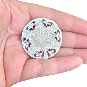 May include: A round, iridescent mother-of-pearl button with a carved floral design. The button features a central rose surrounded by leaves and an openwork border. The button is held in a hand.