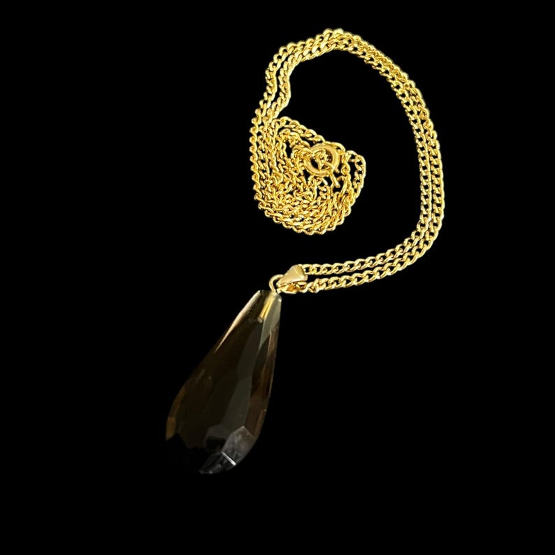 May include: A gold-colored chain necklace with a teardrop-shaped pendant. The pendant is a faceted, dark-colored crystal. The necklace is displayed against a black background.